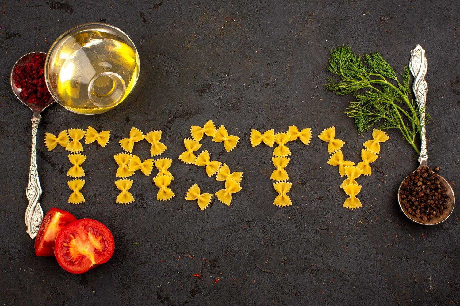 Achieving oil purity: The word 'Tasty' spelled with pasta, surrounded by tomatoes, dill, spices, and a bowl of cooking oil on a dark surface.